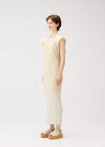 A person with short brown hair stands in a light, minimalist space, smiling gently while wearing the LUCID PLEATS collection from PLEATS PLEASE ISSEY MIYAKE—cream pleated pants, a sleeveless beige tunic, and a sheer long vest.