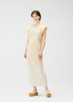 A person with short brown hair stands against a white background, wearing the LUCID PLEATS dress by PLEATS PLEASE ISSEY MIYAKE, styled with a versatile vest and beige platform sandals, looking relaxed and smiling slightly.