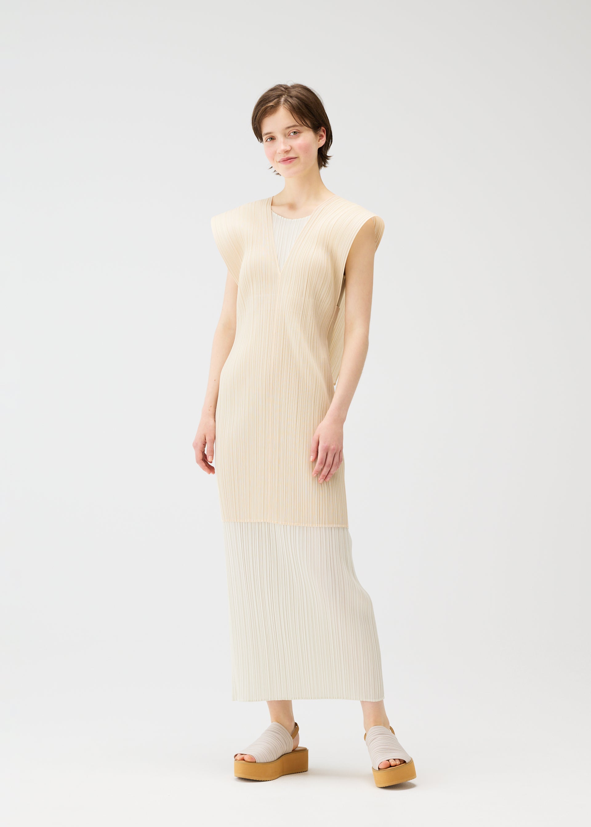 Zoomed view A person with short brown hair stands against a white background, wearing the LUCID PLEATS dress by PLEATS PLEASE ISSEY MIYAKE, styled with a versatile vest and beige platform sandals, looking relaxed and smiling slightly.