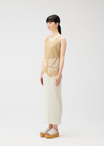 A woman stands in profile against a white background, wearing the I SPY! vest by PLEATS PLEASE ISSEY MIYAKE with a multicolored pattern and a long pleated skirt. She has long black hair in a ponytail and wears platform sandals.