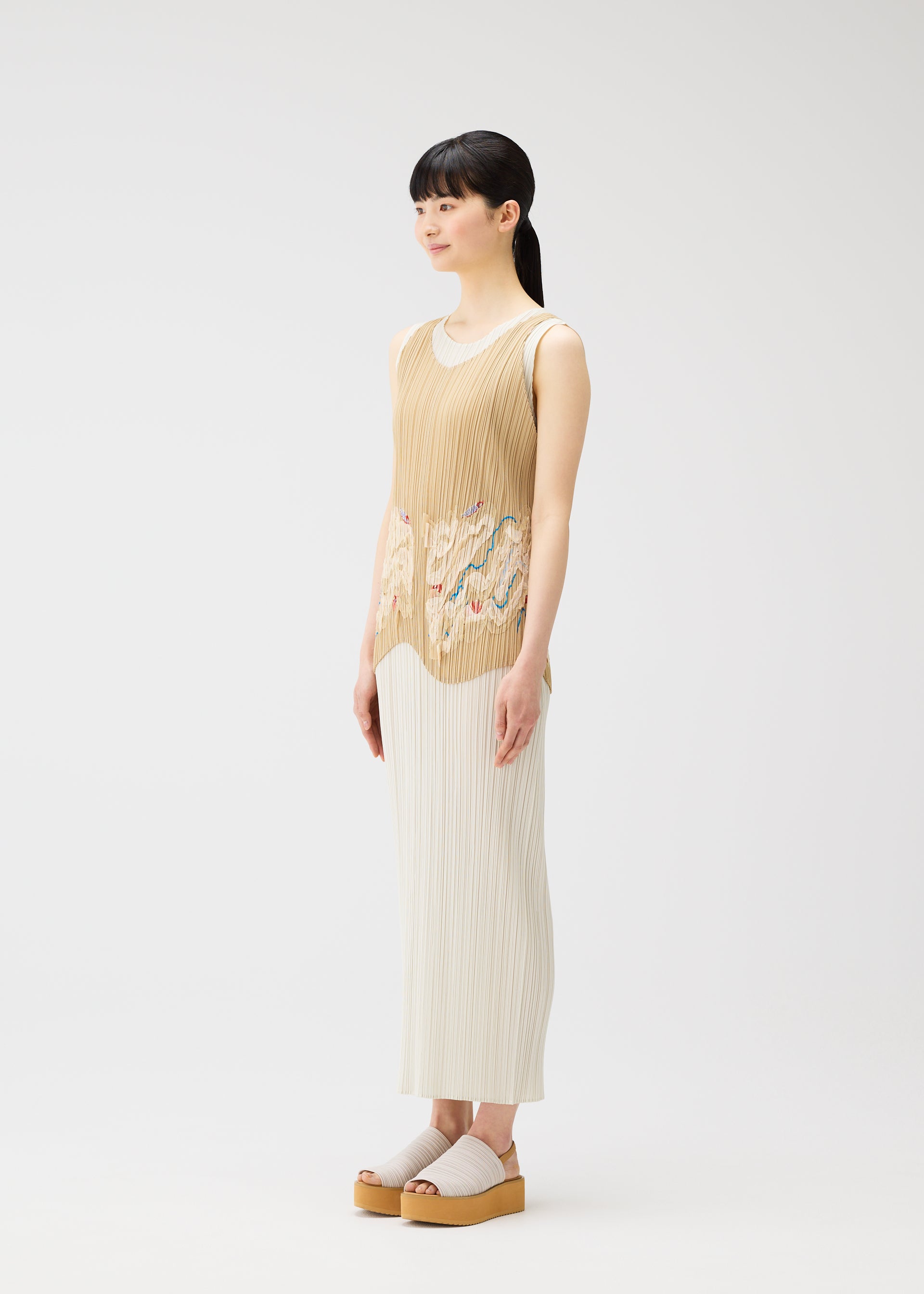 Zoomed view A woman stands in profile against a white background, wearing the I SPY! vest by PLEATS PLEASE ISSEY MIYAKE with a multicolored pattern and a long pleated skirt. She has long black hair in a ponytail and wears platform sandals.