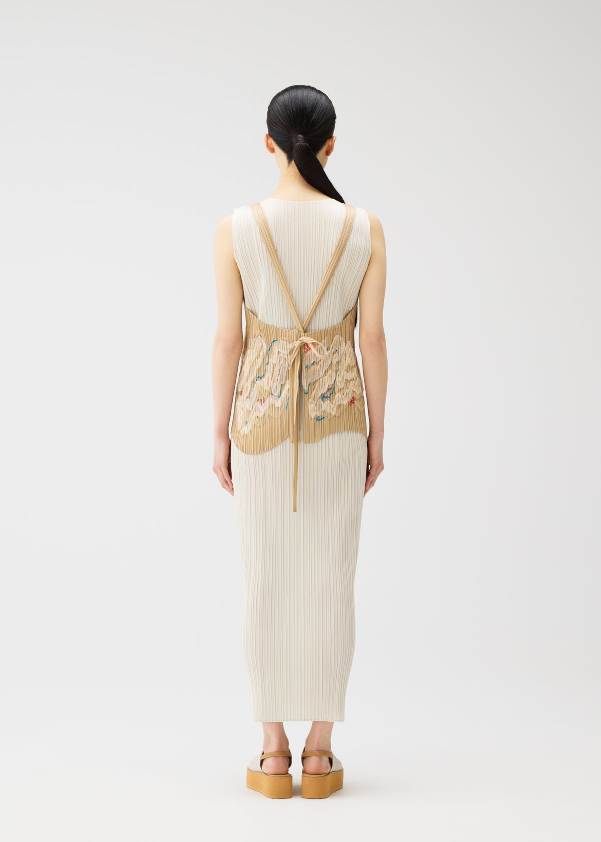 A woman with long dark hair faces away, wearing the PLEATS PLEASE ISSEY MIYAKE "I SPY!" cream ribbed dress and apron-style vest, embroidered with seashells, paired with tan platform sandals. Set against a plain white background. view 9 of 11