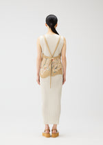 A woman with long dark hair faces away, wearing the PLEATS PLEASE ISSEY MIYAKE "I SPY!" cream ribbed dress and apron-style vest, embroidered with seashells, paired with tan platform sandals. Set against a plain white background.