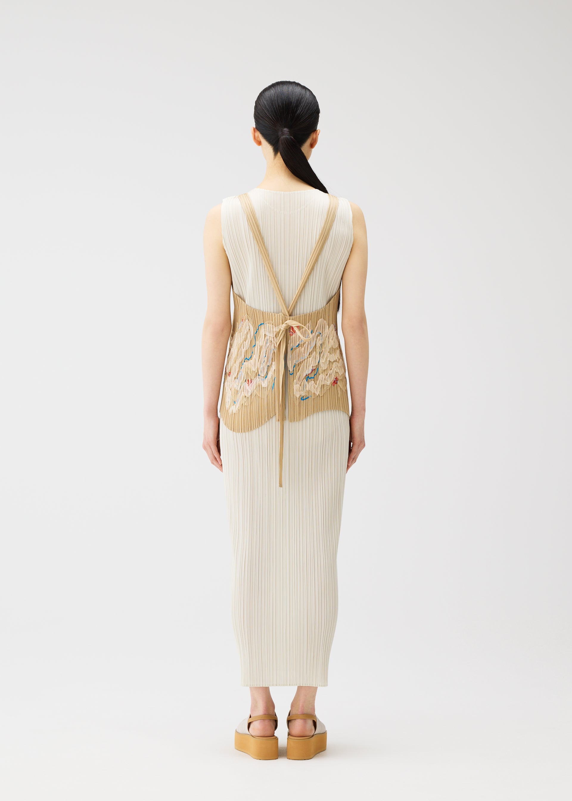 Zoomed view A woman with long dark hair faces away, wearing the PLEATS PLEASE ISSEY MIYAKE "I SPY!" cream ribbed dress and apron-style vest, embroidered with seashells, paired with tan platform sandals. Set against a plain white background.