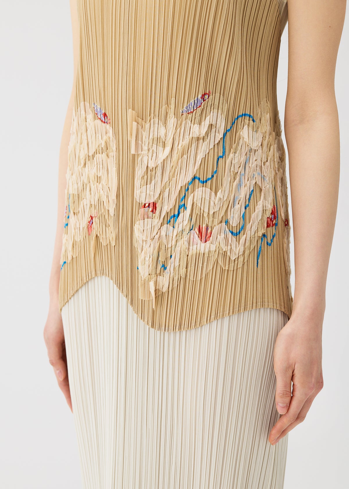 A person wears the I SPY! apron-style vest by PLEATS PLEASE ISSEY MIYAKE—a sleeveless, pleated beige top with abstract cream, blue, and red designs—paired with a white pleated skirt. Image shows torso and upper legs. view 10 of 11