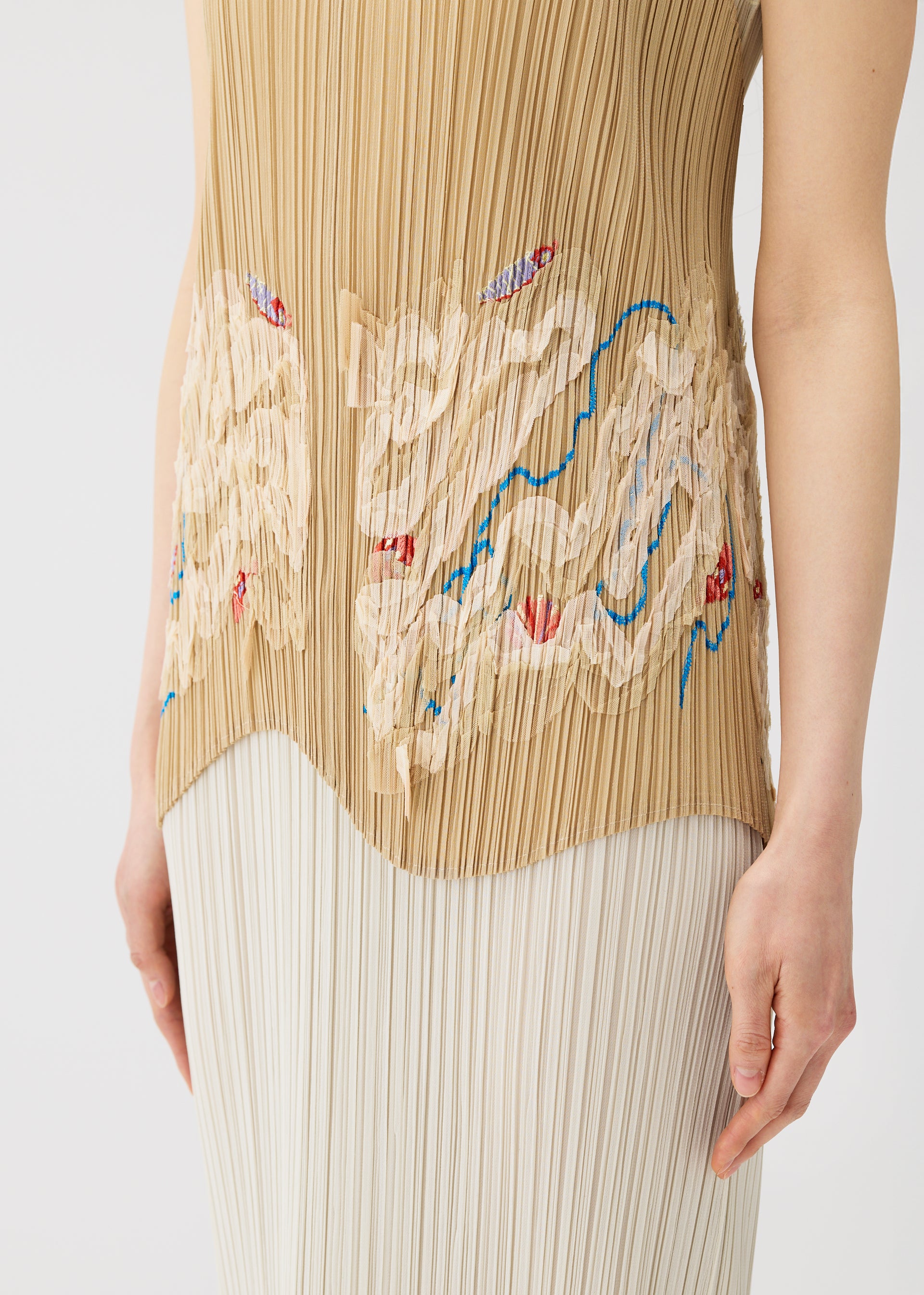 Zoomed view A person wears the I SPY! apron-style vest by PLEATS PLEASE ISSEY MIYAKE—a sleeveless, pleated beige top with abstract cream, blue, and red designs—paired with a white pleated skirt. Image shows torso and upper legs.