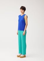 A person with short dark hair smiles in PLEATS PLEASE ISSEY MIYAKE’s STARDUST: a sleeveless blue mesh vest and loose turquoise pants, finished with beige platform sandals, against a plain white background.