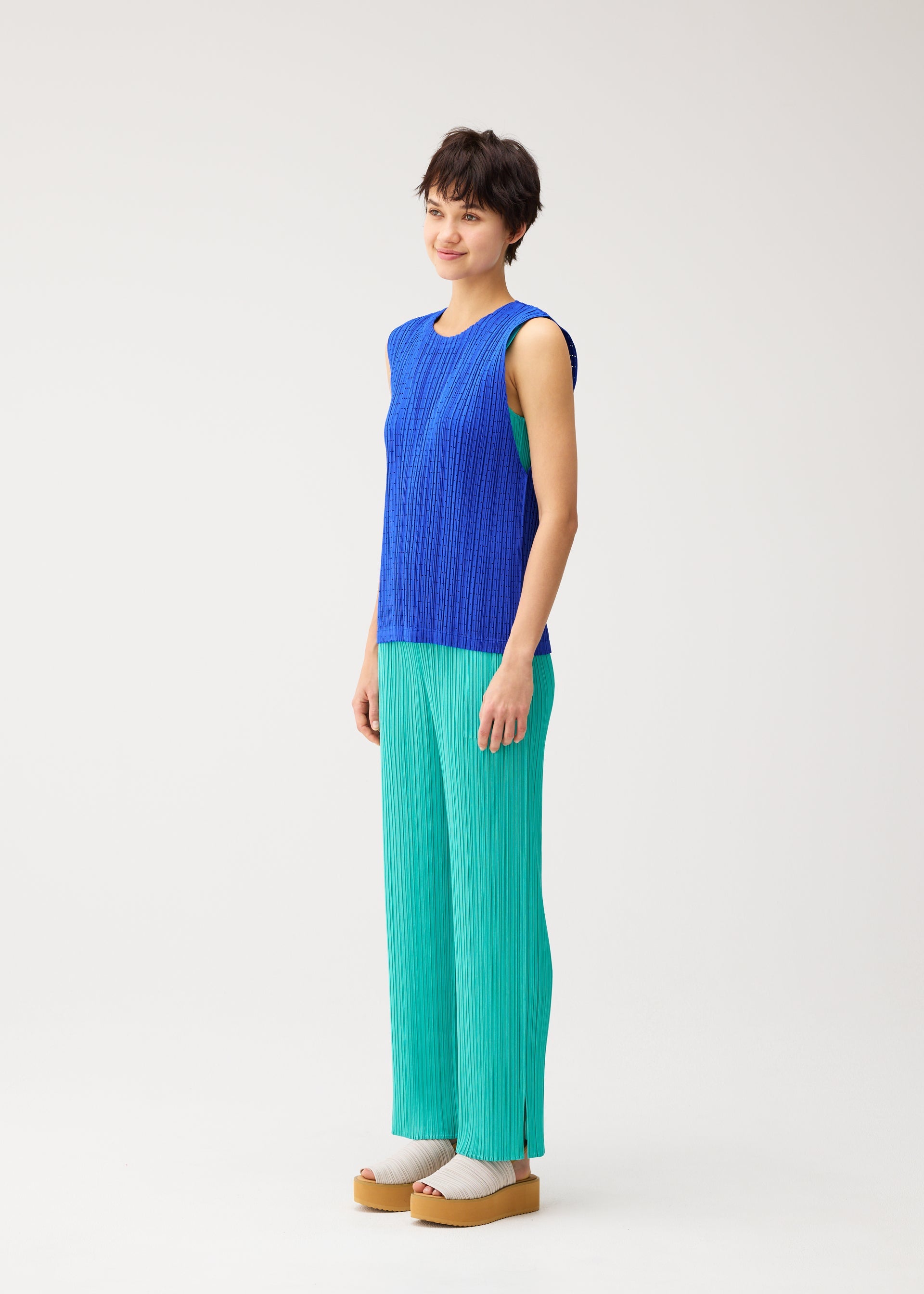 Zoomed view A person with short dark hair smiles in PLEATS PLEASE ISSEY MIYAKE’s STARDUST: a sleeveless blue mesh vest and loose turquoise pants, finished with beige platform sandals, against a plain white background.