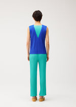 A person with short dark hair stands against a plain light background wearing the STARDUST sleeveless blue top with a deep V back and matte finish, and voluminous pleated turquoise pants by PLEATS PLEASE ISSEY MIYAKE, paired with tan platform shoes.