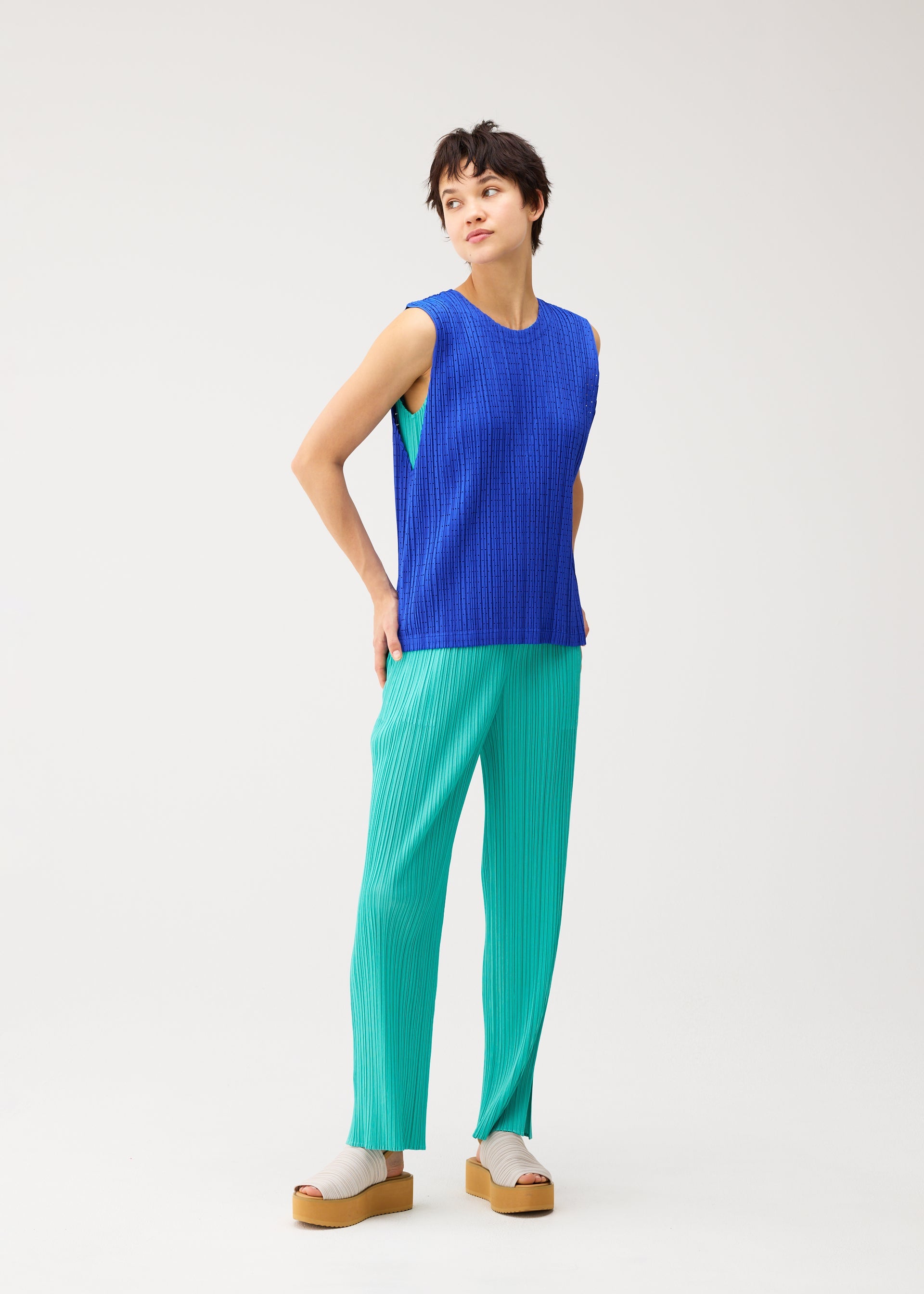 Zoomed view A person stands against a plain white background, wearing the STARDUST sleeveless blue top by PLEATS PLEASE ISSEY MIYAKE, paired with loose turquoise pants and beige platform sandals. They look slightly to the side with hands behind their back.