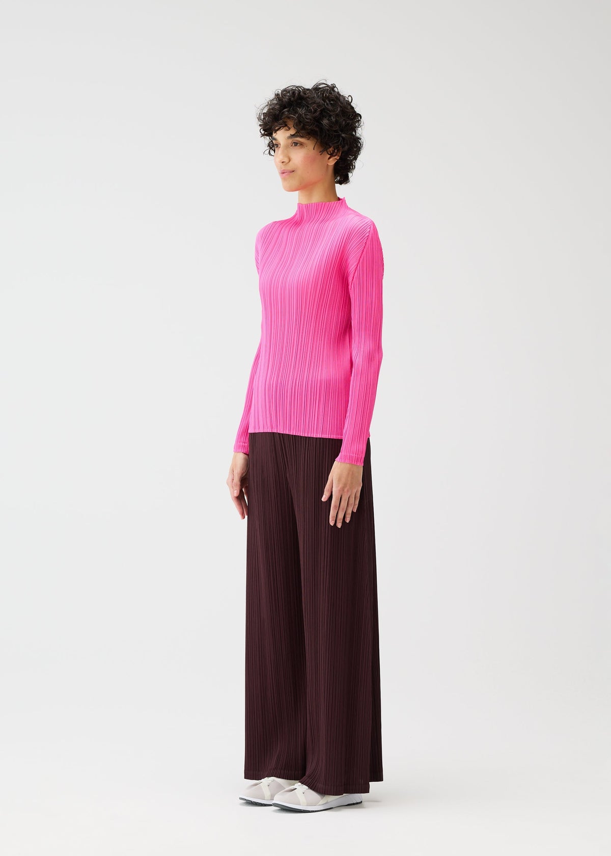 A person with short curly hair wears the MONTHLY COLORS : JANUARY top by PLEATS PLEASE ISSEY MIYAKE, paired with mid-weight dark brown straight pants with pockets and white shoes, standing against a plain white background. view 7 of 8