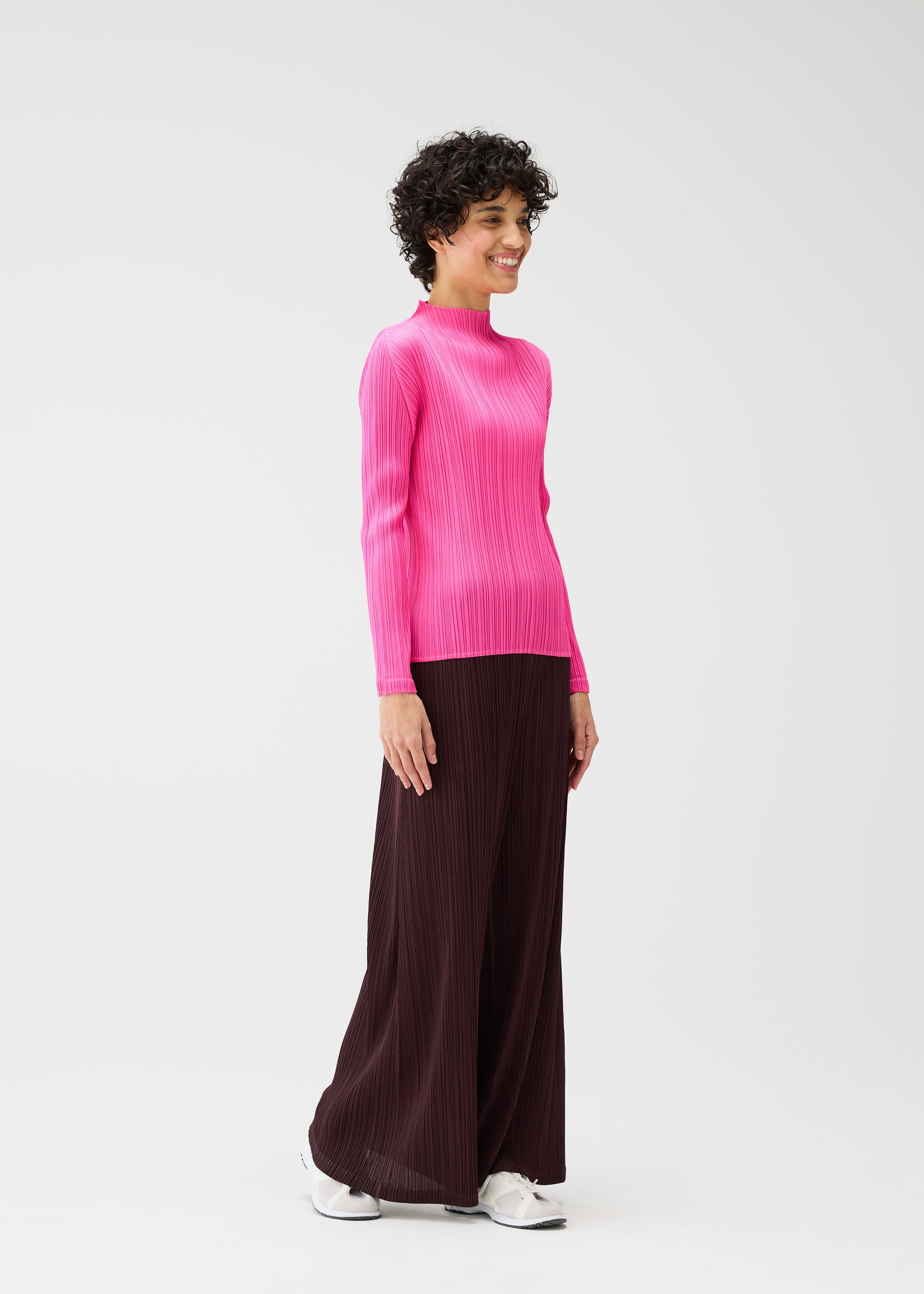 Zoomed view A person stands smiling against a plain white background, wearing the MONTHLY COLORS : JANUARY bright pink long-sleeve top and dark brown mid-weight straight pants with pockets by PLEATS PLEASE ISSEY MIYAKE, paired with white sneakers.