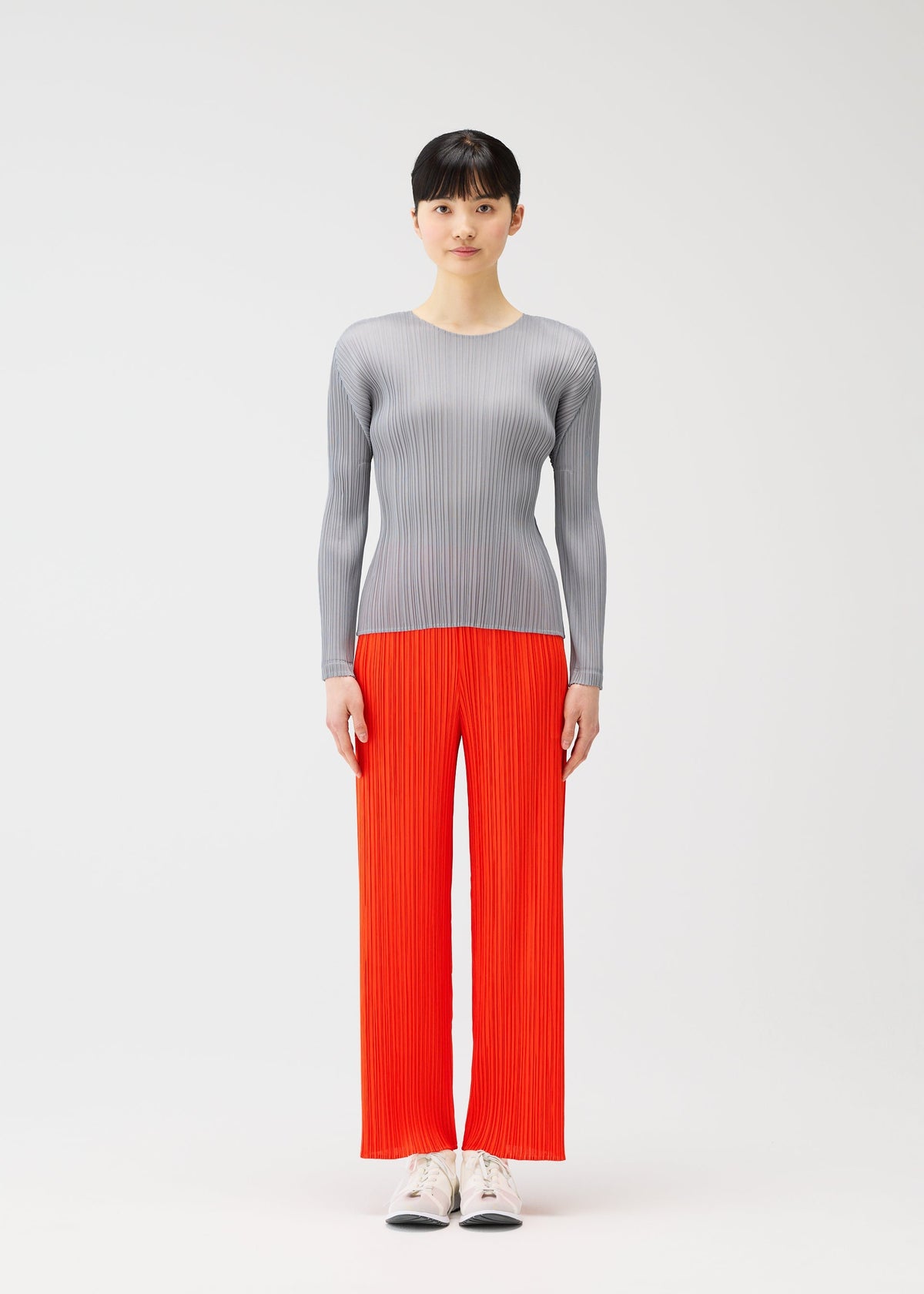 A person stands facing forward against a plain white background, wearing a fitted long-sleeve light gray top and bright orange straight pants from the PLEATS PLEASE ISSEY MIYAKE MONTHLY COLORS : FEBRUARY collection, paired with white shoes. view 7 of 9