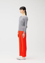 A woman with long dark hair in a ponytail stands in profile against a light background, styled for PLEATS PLEASE ISSEY MIYAKE's MONTHLY COLORS : FEBRUARY in a gray ribbed top, bright orange pants, and white shoes.