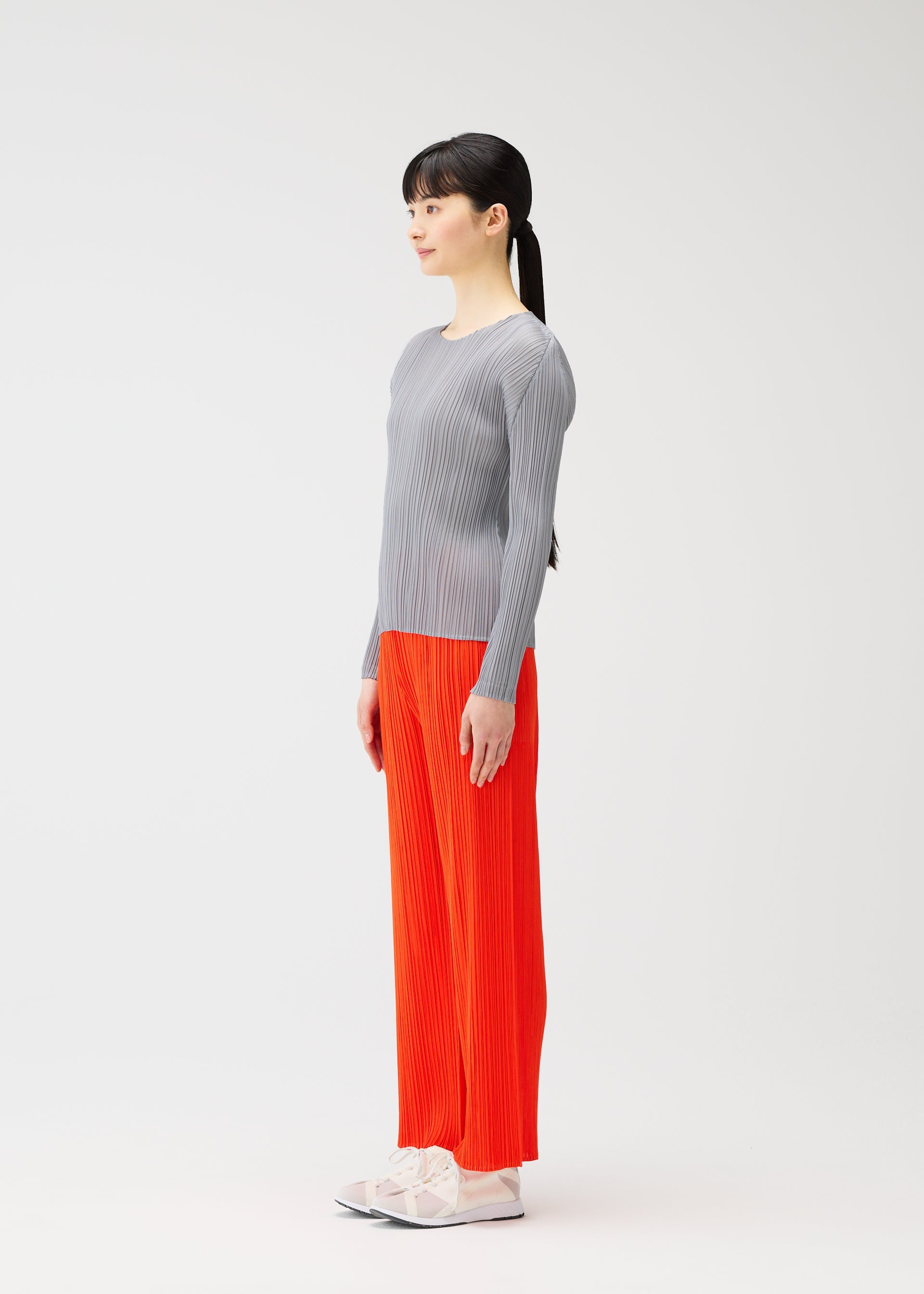 Zoomed view A woman with long dark hair in a ponytail stands in profile against a light background, styled for PLEATS PLEASE ISSEY MIYAKE's MONTHLY COLORS : FEBRUARY in a gray ribbed top, bright orange pants, and white shoes.
