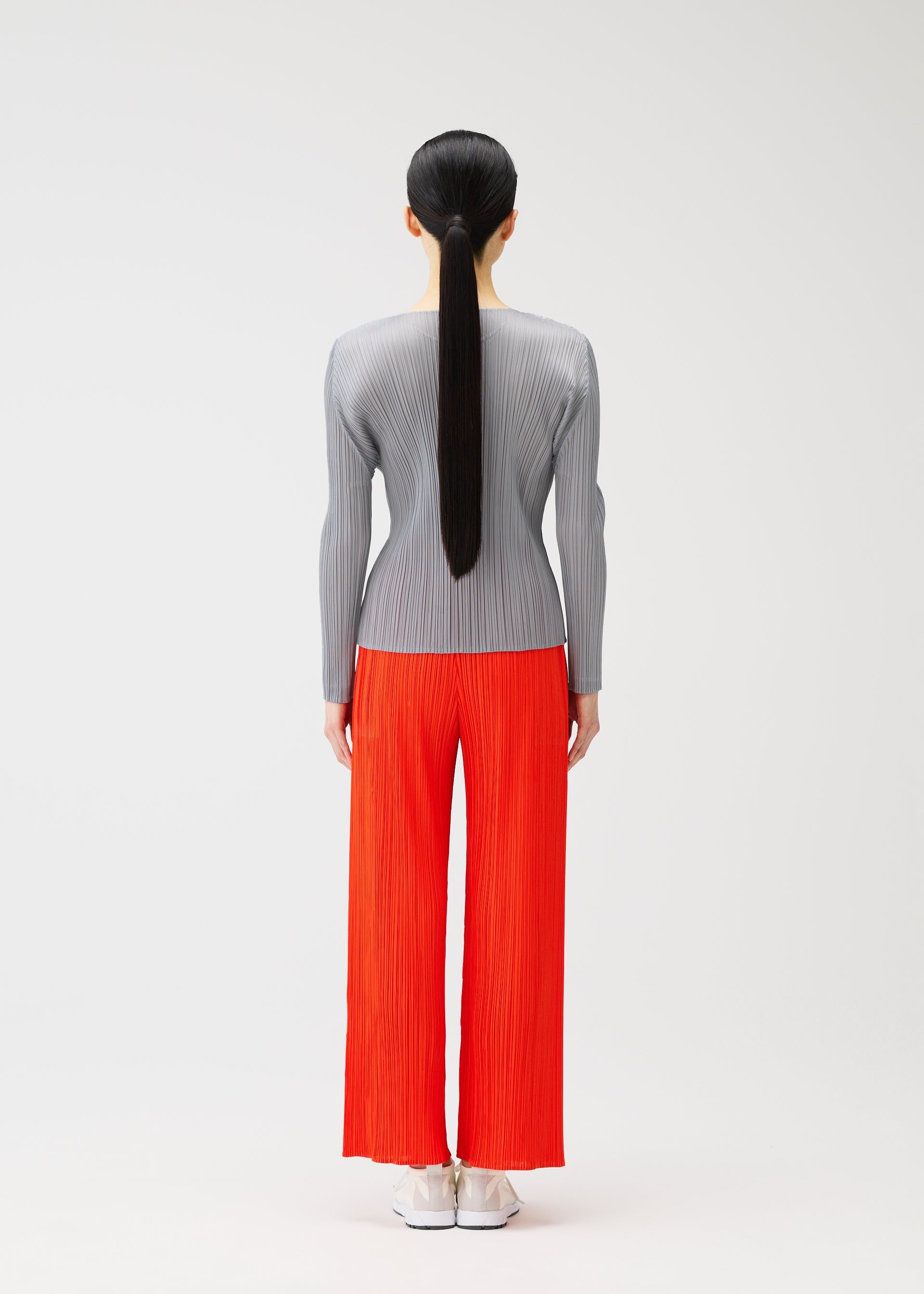 Zoomed view A person with long, straight black hair in a ponytail stands facing away, wearing a gray long-sleeve top and bright red pleated wide-leg pants from the PLEATS PLEASE ISSEY MIYAKE MONTHLY COLORS : FEBRUARY collection, with white shoes and a plain background.