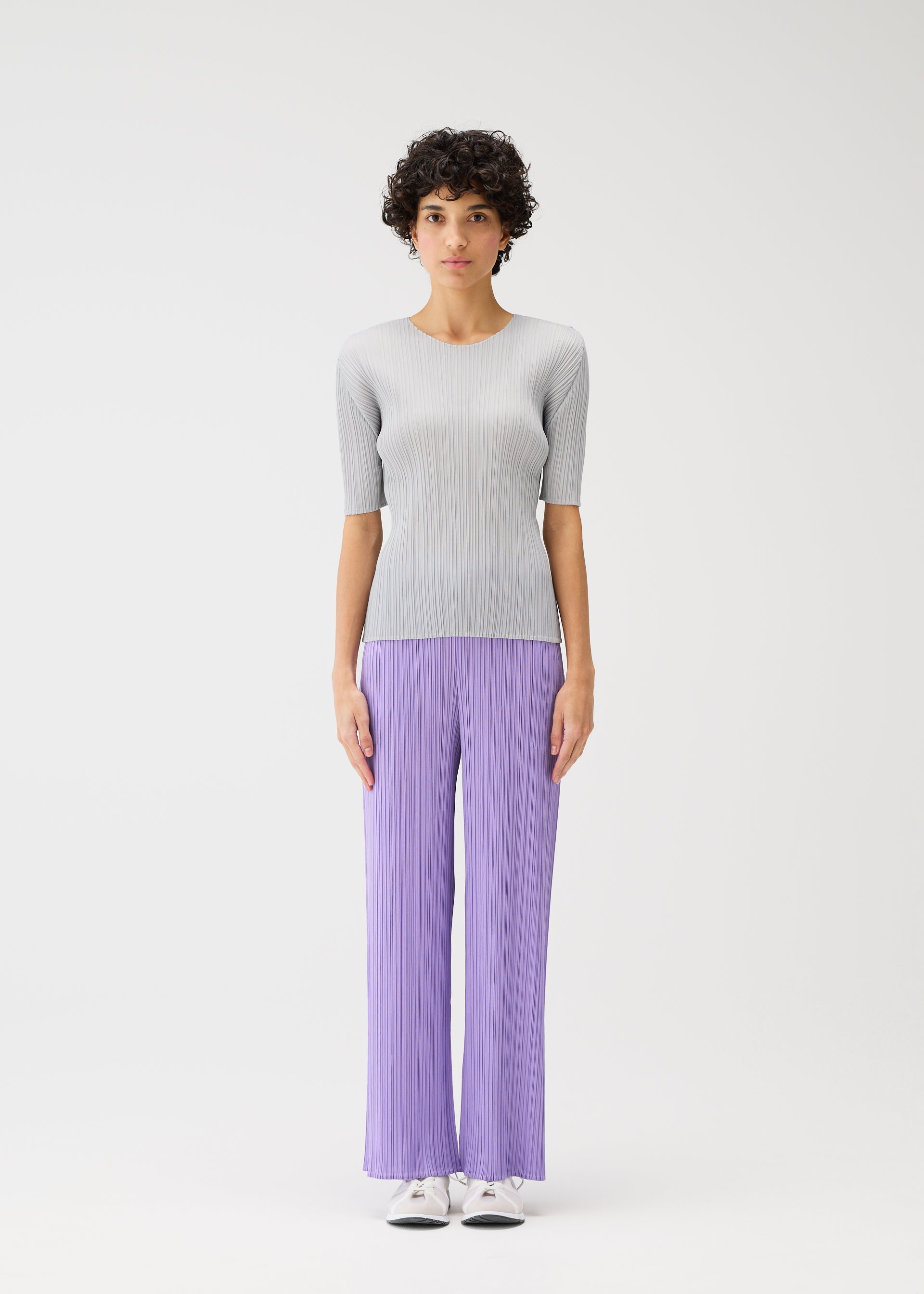 Zoomed view A person stands facing forward against a plain white background, wearing light colors from the MONTHLY COLORS : APRIL collection by PLEATS PLEASE ISSEY MIYAKE—ribbed top, loose straight pants, and white sneakers with simple silhouettes.