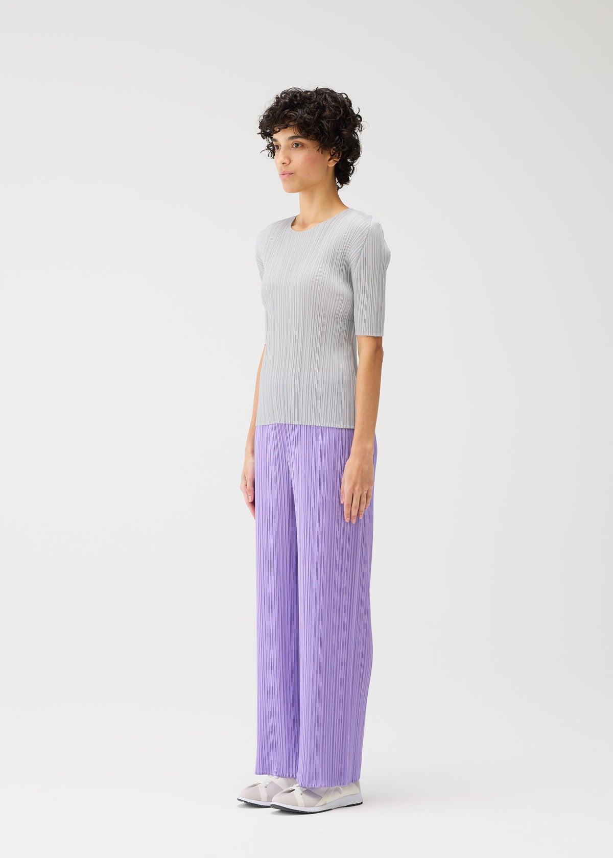 A person with short curly hair stands sideways in a light gray top and lavender loose straight pants from the MONTHLY COLORS : APRIL collection by PLEATS PLEASE ISSEY MIYAKE, paired with white shoes against a plain white background. view 7 of 8