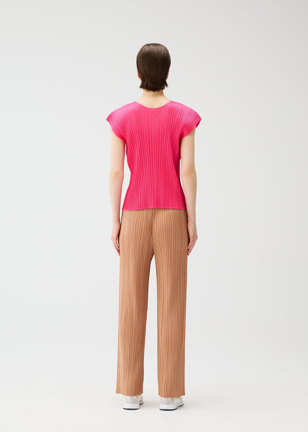 A person stands facing away, wearing the MONTHLY COLORS : JUNE pink pleated top and loose tan pants with side pockets from PLEATS PLEASE ISSEY MIYAKE, paired with white shoes, against a plain white background. view 9 of 10