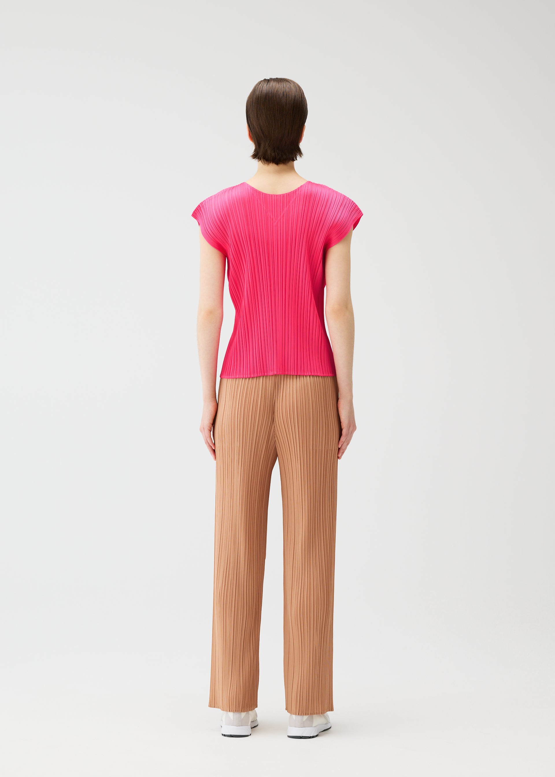 Zoomed view A person stands facing away, wearing the MONTHLY COLORS : JUNE pink pleated top and loose tan pants with side pockets from PLEATS PLEASE ISSEY MIYAKE, paired with white shoes, against a plain white background.