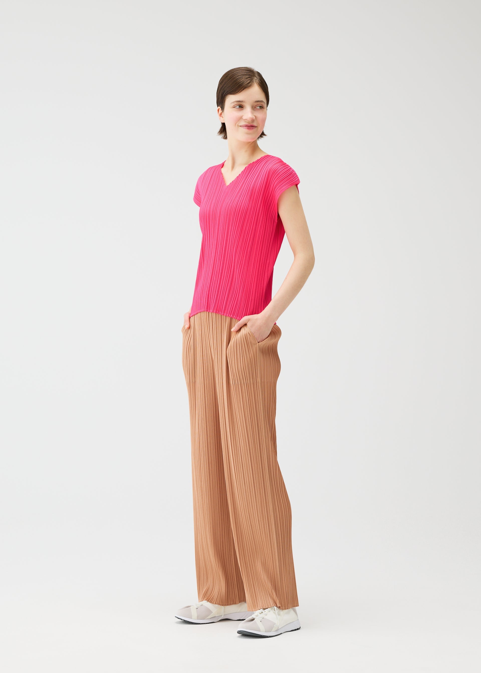 Zoomed view Wearing the MONTHLY COLORS : JUNE bright pink top and loose beige pants from PLEATS PLEASE ISSEY MIYAKE, a person stands against a white background, hands in pockets, looking to the side with a slight smile and white sneakers.