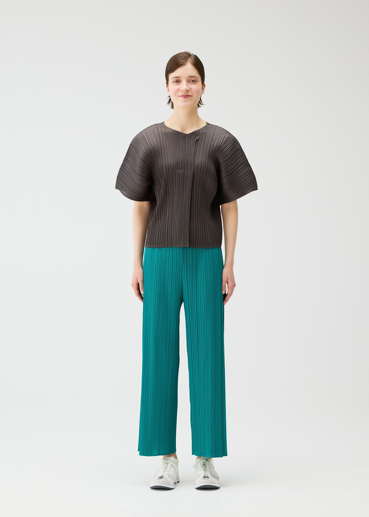 A person with short brown hair and a neutral expression wears the PLEATS PLEASE ISSEY MIYAKE MONTHLY COLORS: JUNE wide-sleeved textured black top, coastal-colored tucked pants with subtle pockets, and white sneakers, against a plain white background. view 7 of 10