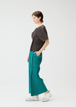 A woman stands in profile against a white background, wearing PLEATS PLEASE ISSEY MIYAKE MONTHLY COLORS : JUNE—a short-sleeved pleated top and teal wide-leg pants. Her left hand is in her pocket, and she smiles gently, finished with crisp white sneakers.