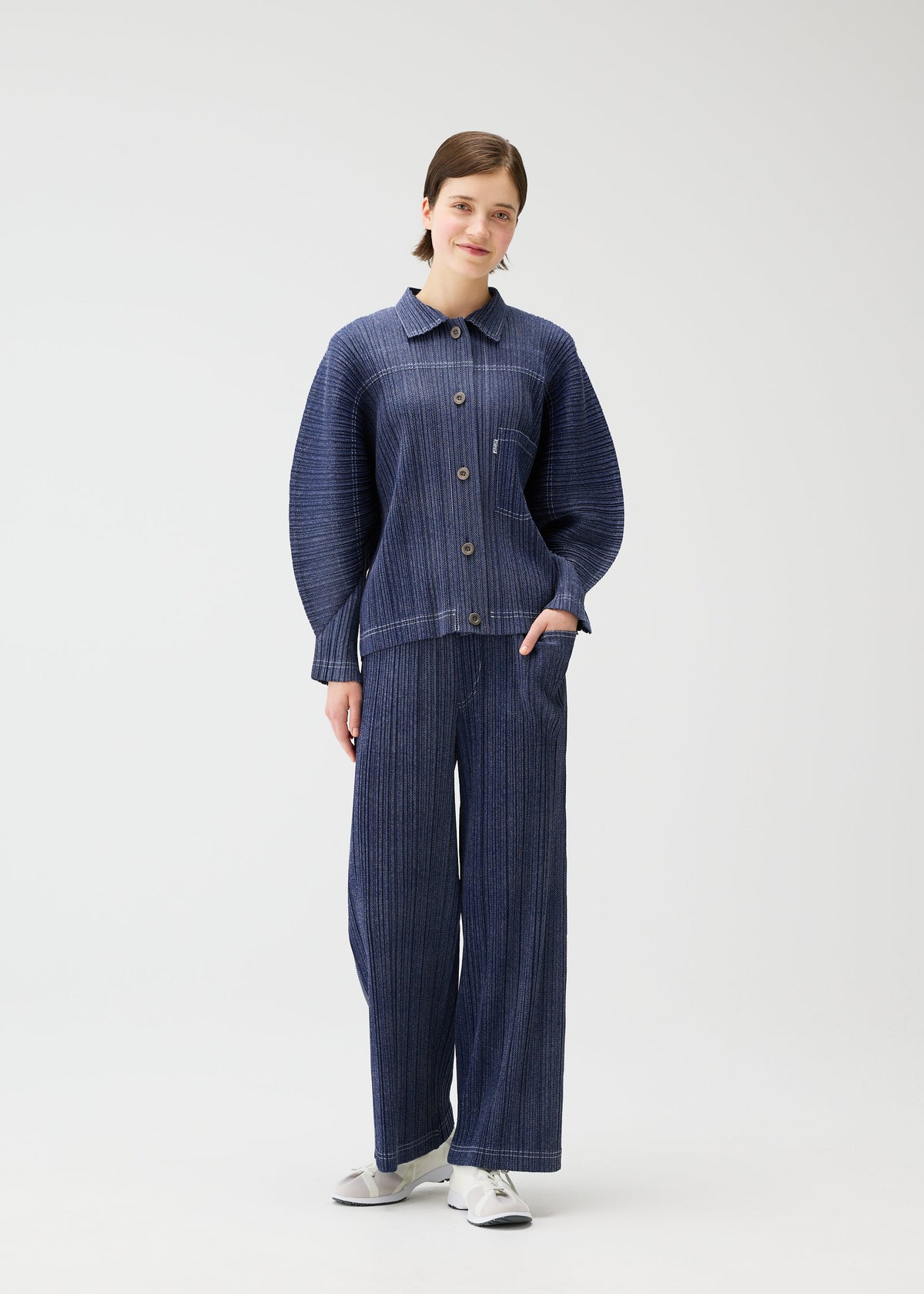 A woman stands against a white background wearing the COTTON DENIM set by PLEATS PLEASE ISSEY MIYAKE: a loose-fitting, dark blue pinstriped jacket and matching wide pants with white sneakers. She smiles slightly with one hand in her pocket. view 8 of 9