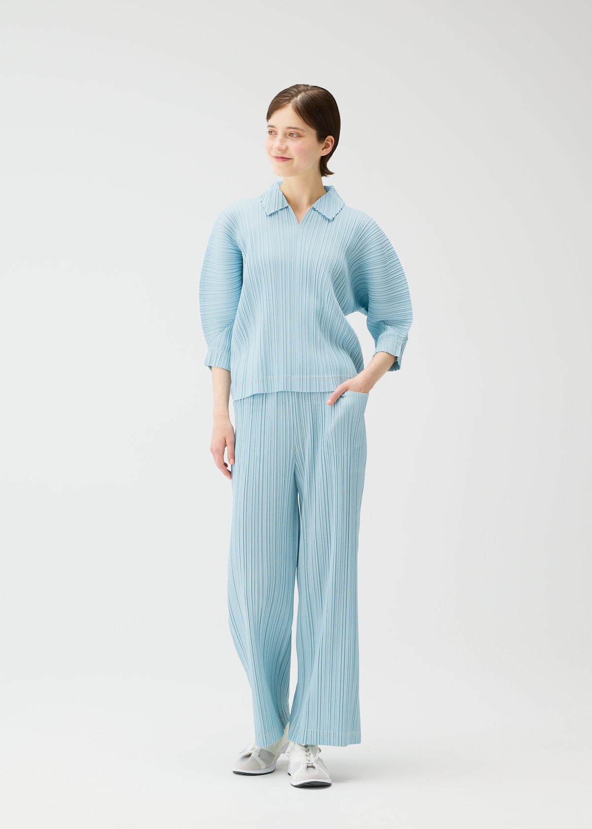 A person stands against a white background wearing the PLEATS PLEASE ISSEY MIYAKE COTTON DENIM—a light blue, cotton-blend pleated top and matching wide pants—with white sneakers. One hand is in their pocket as they smile, looking slightly to the side. view 9 of 9