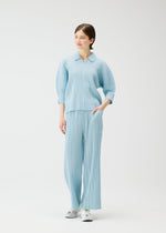 A person stands against a white background wearing the PLEATS PLEASE ISSEY MIYAKE COTTON DENIM—a light blue, cotton-blend pleated top and matching wide pants—with white sneakers. One hand is in their pocket as they smile, looking slightly to the side.
