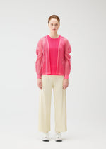 A woman faces forward in a bright pink mesh jacket over a pink top, styled with cream OPAQUE BOTTOMS from PLEATS PLEASE ISSEY MIYAKE and white sneakers, set against a plain white background.