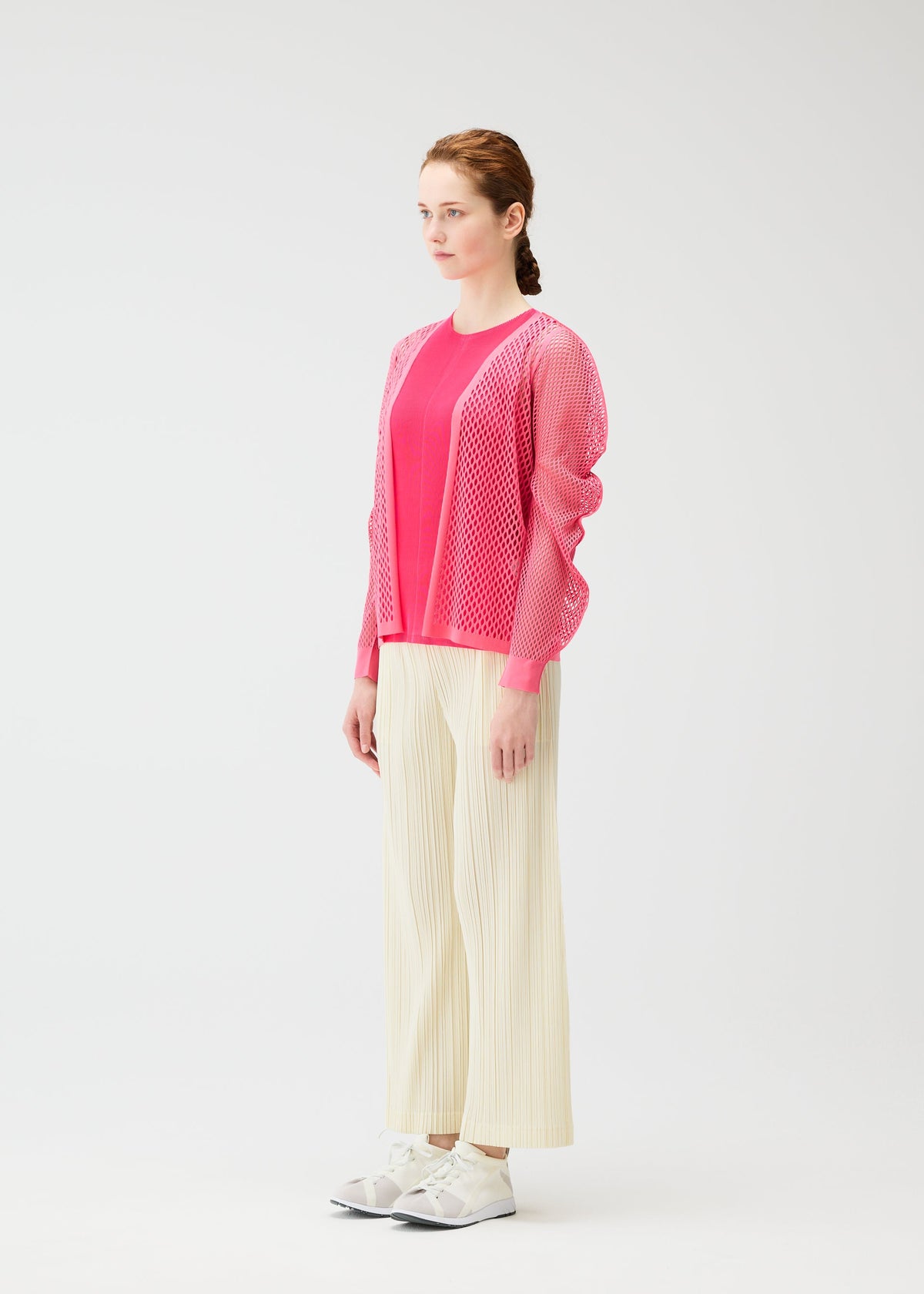A woman with braided hair in profile stands against a white background, wearing a bright pink mesh cardigan, a pink top, OPAQUE BOTTOMS by PLEATS PLEASE ISSEY MIYAKE, and white sneakers. view 6 of 8