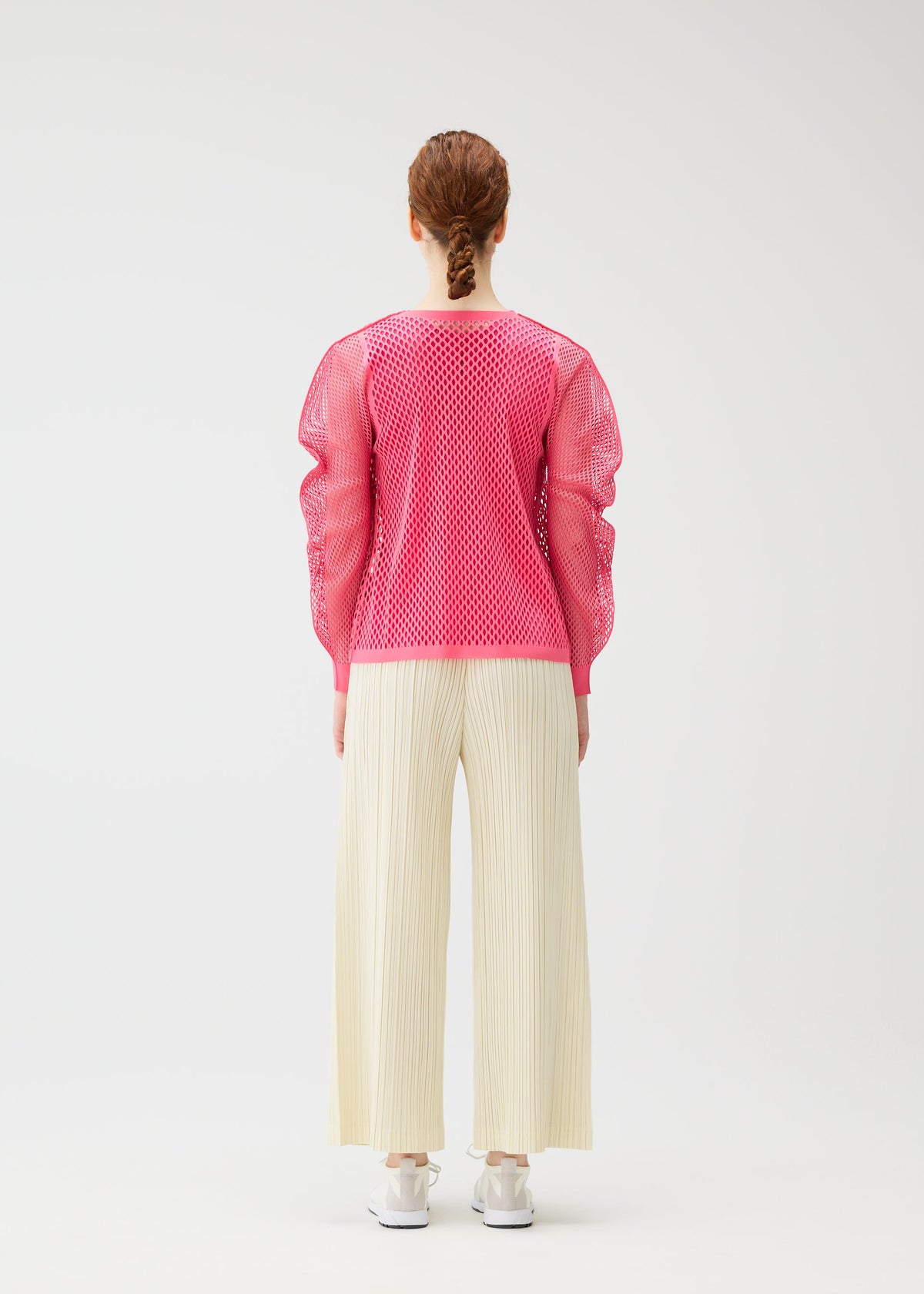 A person with braided hair faces away, wearing a bright pink matte-texture mesh top, white shoes, and cream OPAQUE BOTTOMS by PLEATS PLEASE ISSEY MIYAKE against a plain white background. view 7 of 8