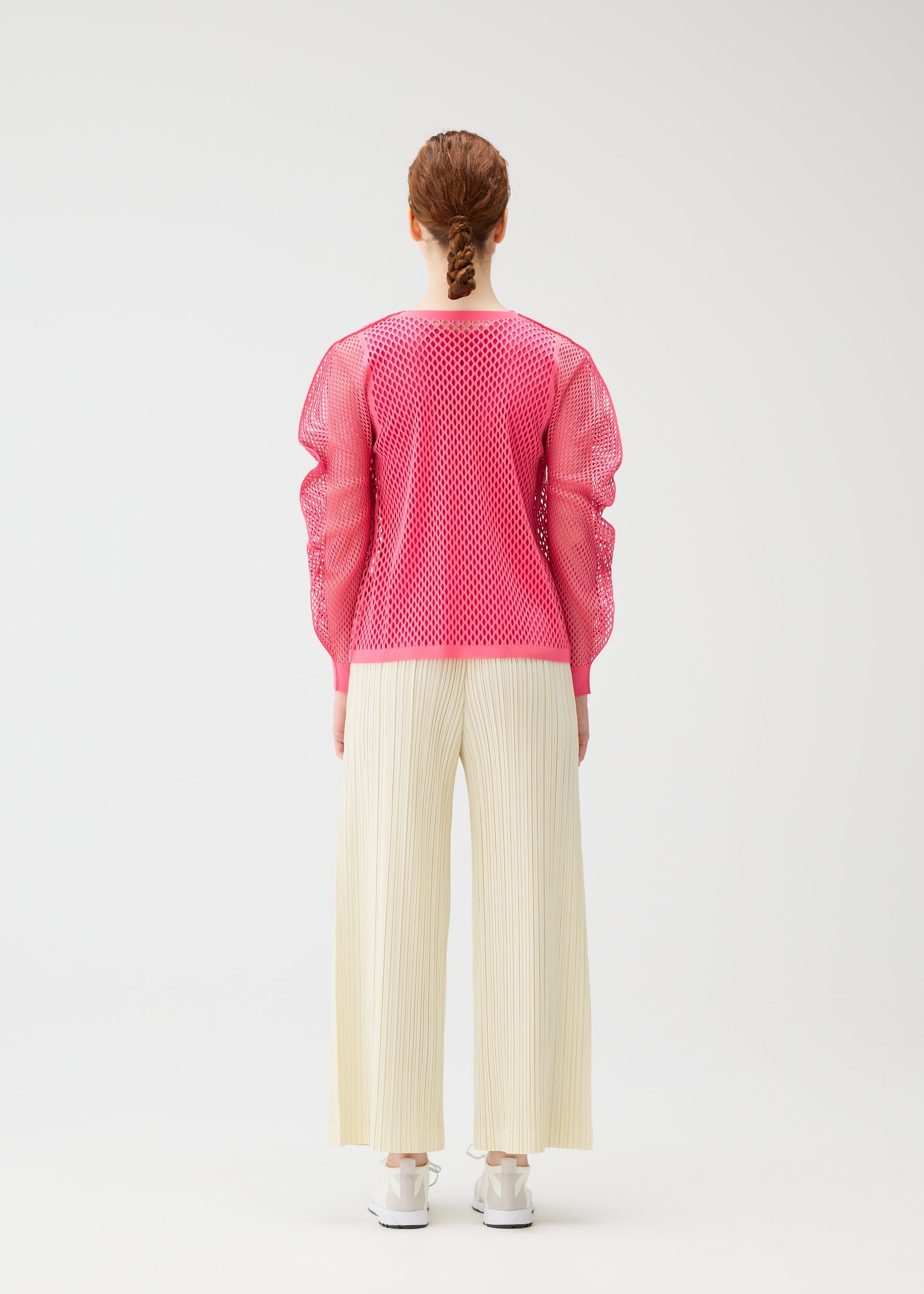 Zoomed view A person with braided hair faces away, wearing a bright pink matte-texture mesh top, white shoes, and cream OPAQUE BOTTOMS by PLEATS PLEASE ISSEY MIYAKE against a plain white background.