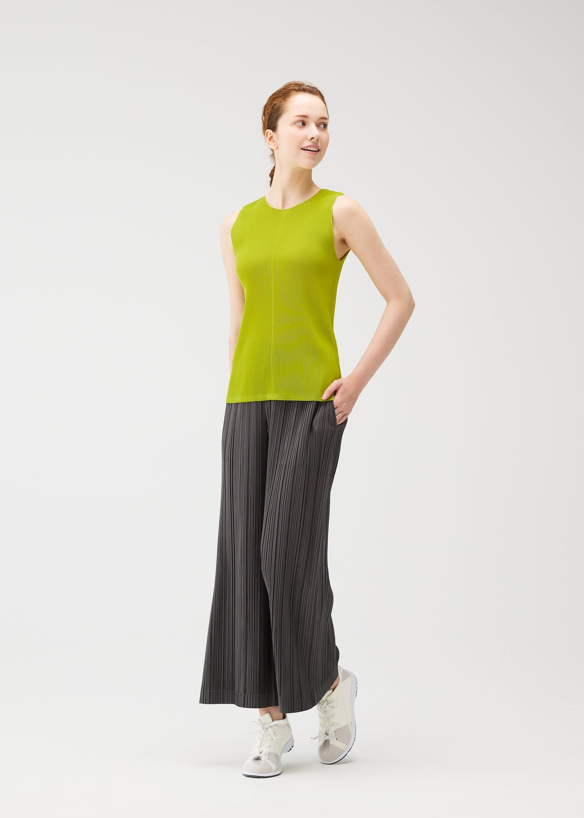 Wearing OPAQUE BOTTOMS by PLEATS PLEASE ISSEY MIYAKE, a person stands against a white background, paired with a sleeveless green top and white sneakers, smiling and looking to the side with hands in their pockets. view 8 of 8