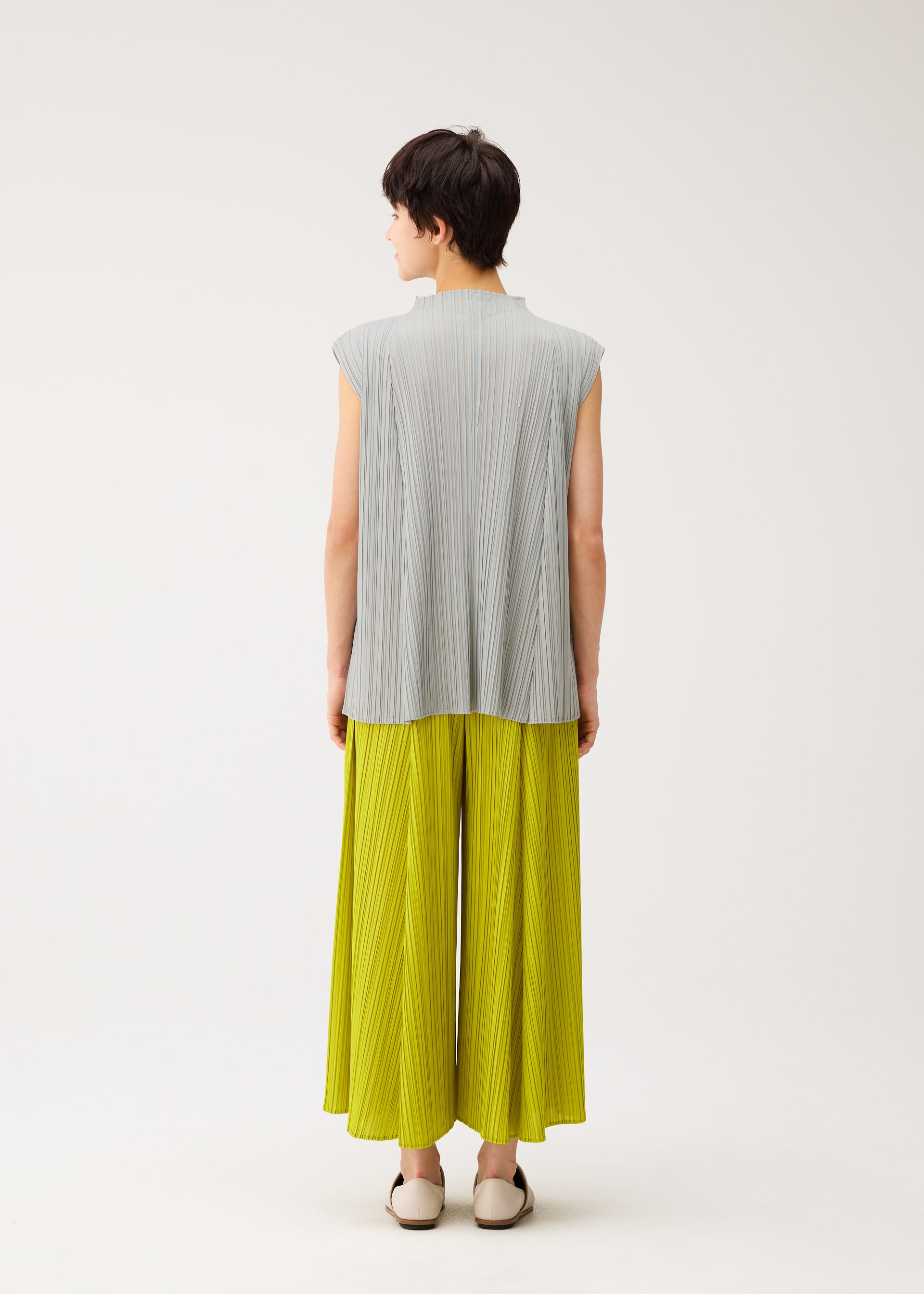 Zoomed view A person with short dark hair faces away, wearing the MELLOW PLEATS sleeveless light gray top and bright yellow wide-legged pants by PLEATS PLEASE ISSEY MIYAKE, paired with white flats against a plain light background.