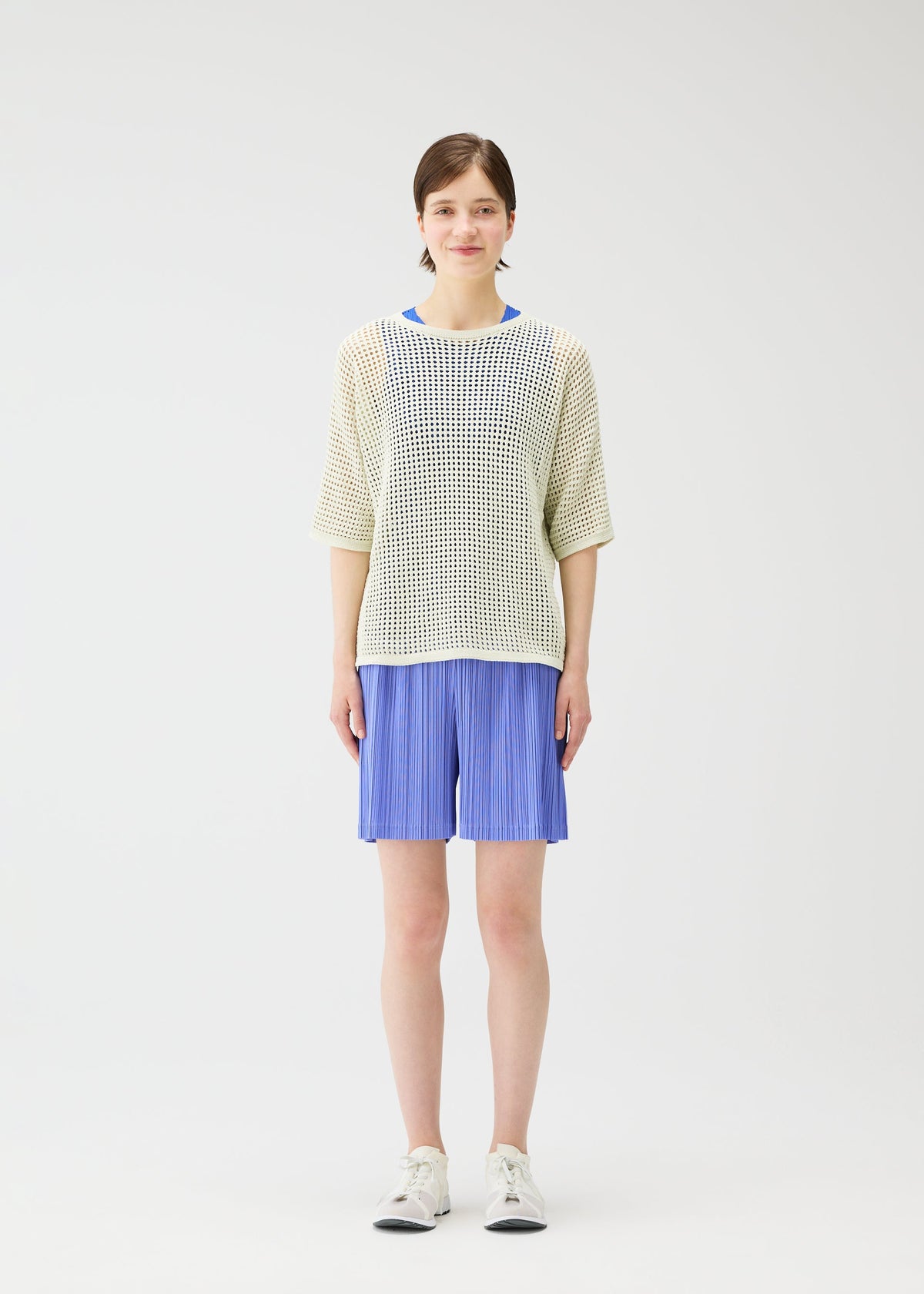 A woman faces forward against a white background, wearing PLEATS PLEASE ISSEY MIYAKE LOOP BOTTOMS in light blue with pockets, paired with a loose-knit white sweater over a blue top and white sneakers. view 5 of 8