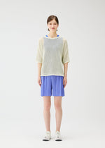 A woman faces forward against a white background, wearing PLEATS PLEASE ISSEY MIYAKE LOOP BOTTOMS in light blue with pockets, paired with a loose-knit white sweater over a blue top and white sneakers.