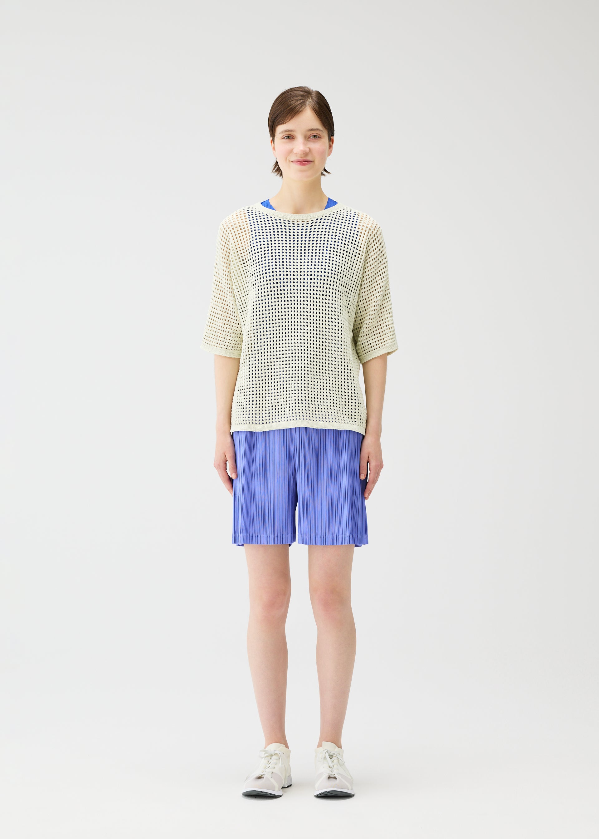 Zoomed view A woman faces forward against a white background, wearing PLEATS PLEASE ISSEY MIYAKE LOOP BOTTOMS in light blue with pockets, paired with a loose-knit white sweater over a blue top and white sneakers.