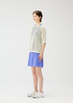 A person with short brown hair stands against a white background, smiling softly while wearing PLEATS PLEASE ISSEY MIYAKE LOOP BOTTOMS—blue pleated shorts—paired with a mesh cream top, blue shirt, and white athletic shoes.