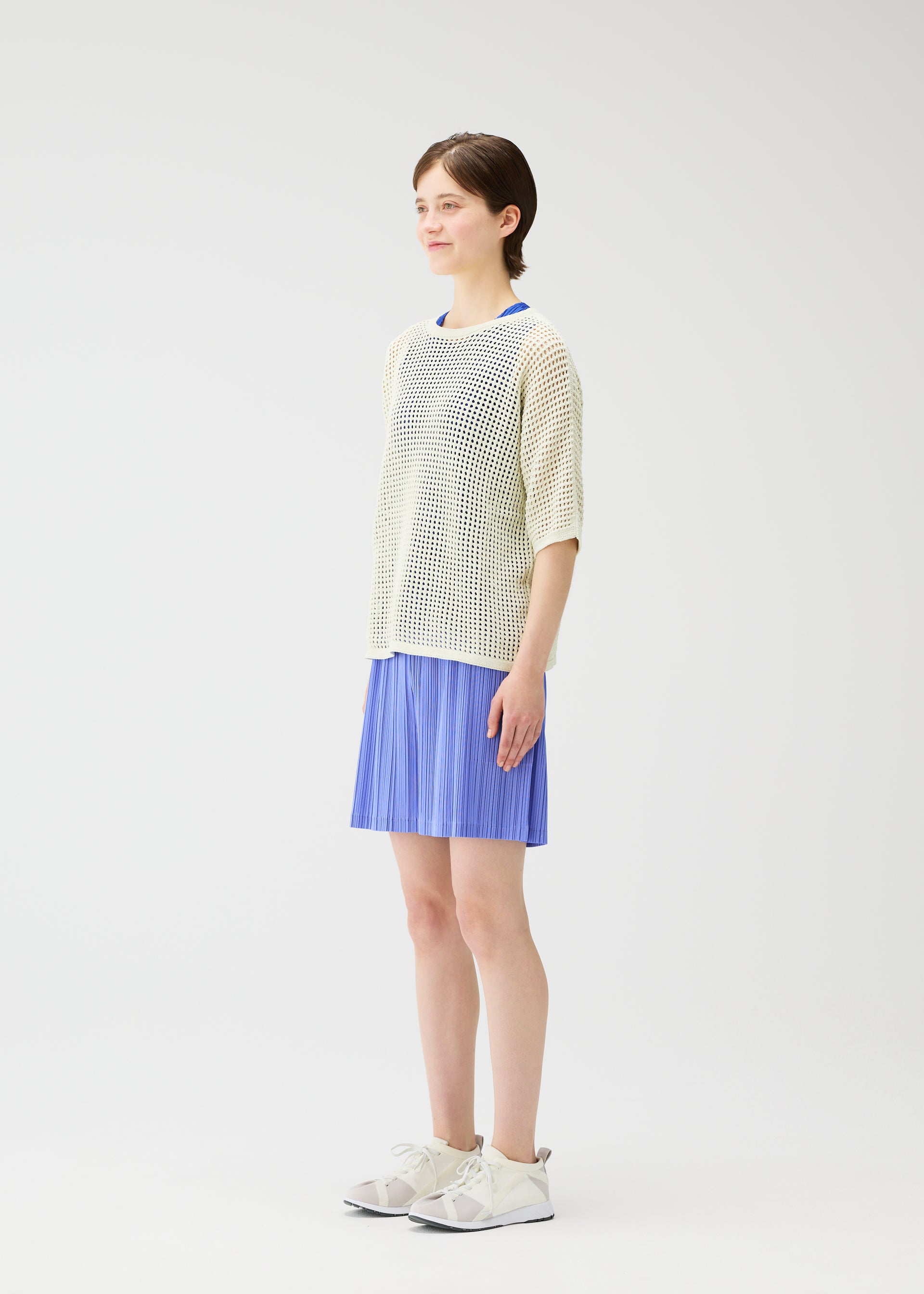 Zoomed view A person with short brown hair stands against a white background, smiling softly while wearing PLEATS PLEASE ISSEY MIYAKE LOOP BOTTOMS—blue pleated shorts—paired with a mesh cream top, blue shirt, and white athletic shoes.