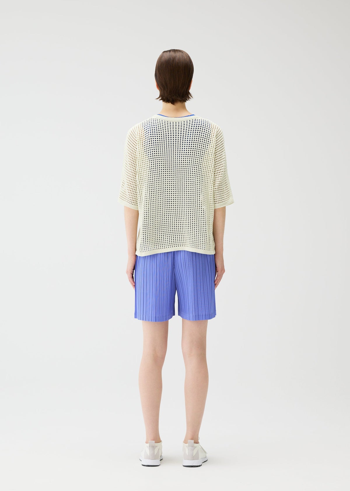A person stands facing away in a loose white mesh top, LOOP BOTTOMS light blue recycled polyester shorts with pockets by PLEATS PLEASE ISSEY MIYAKE, and white shoes against a plain white background. view 7 of 8
