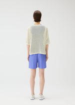 A person stands facing away in a loose white mesh top, LOOP BOTTOMS light blue recycled polyester shorts with pockets by PLEATS PLEASE ISSEY MIYAKE, and white shoes against a plain white background.