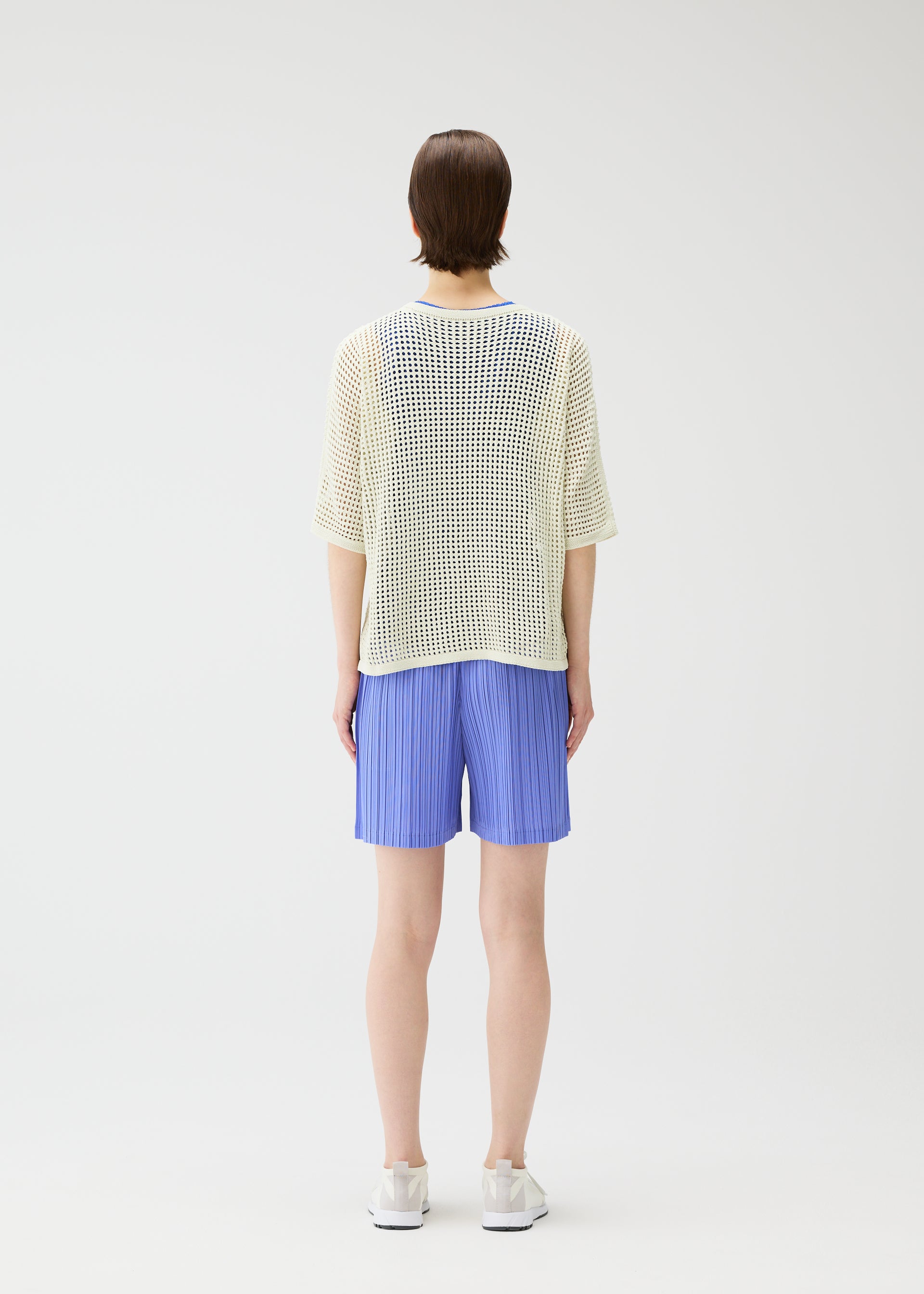 Zoomed view A person stands facing away in a loose white mesh top, LOOP BOTTOMS light blue recycled polyester shorts with pockets by PLEATS PLEASE ISSEY MIYAKE, and white shoes against a plain white background.