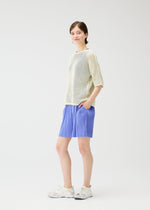 A person stands against a white background, hands in pockets and smiling slightly, wearing a light mesh sweater, LOOP BOTTOMS blue shorts by PLEATS PLEASE ISSEY MIYAKE with pockets, and white sneakers.