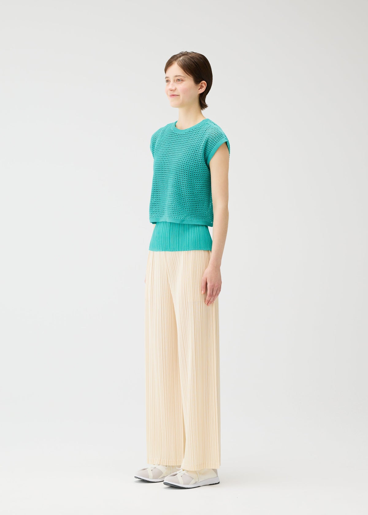 A person stands against a white background wearing cream LOOP BOTTOMS by PLEATS PLEASE ISSEY MIYAKE, a turquoise short-sleeve knit top, and white shoes. They face slightly right and smile softly. view 6 of 8