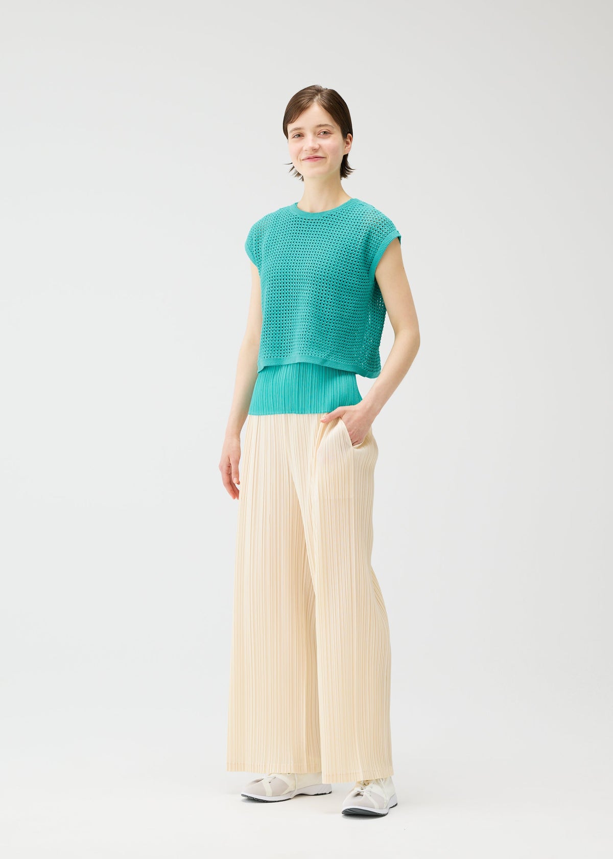 A person stands against a plain white background, wearing a teal textured short-sleeve top, PLEATS PLEASE ISSEY MIYAKE LOOP BOTTOMS in pleated cream, and white sneakers, with one hand in their pocket and a slight smile. view 8 of 8