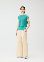 A person stands against a plain white background, wearing a teal textured short-sleeve top, PLEATS PLEASE ISSEY MIYAKE LOOP BOTTOMS in pleated cream, and white sneakers, with one hand in their pocket and a slight smile.