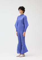 A person with short, curly hair stands against a plain background, wearing the SKYLINE set by PLEATS PLEASE ISSEY MIYAKE—blue pleated long-sleeve top with wide sleeves and matching city-inspired pants—paired with light-colored shoes.