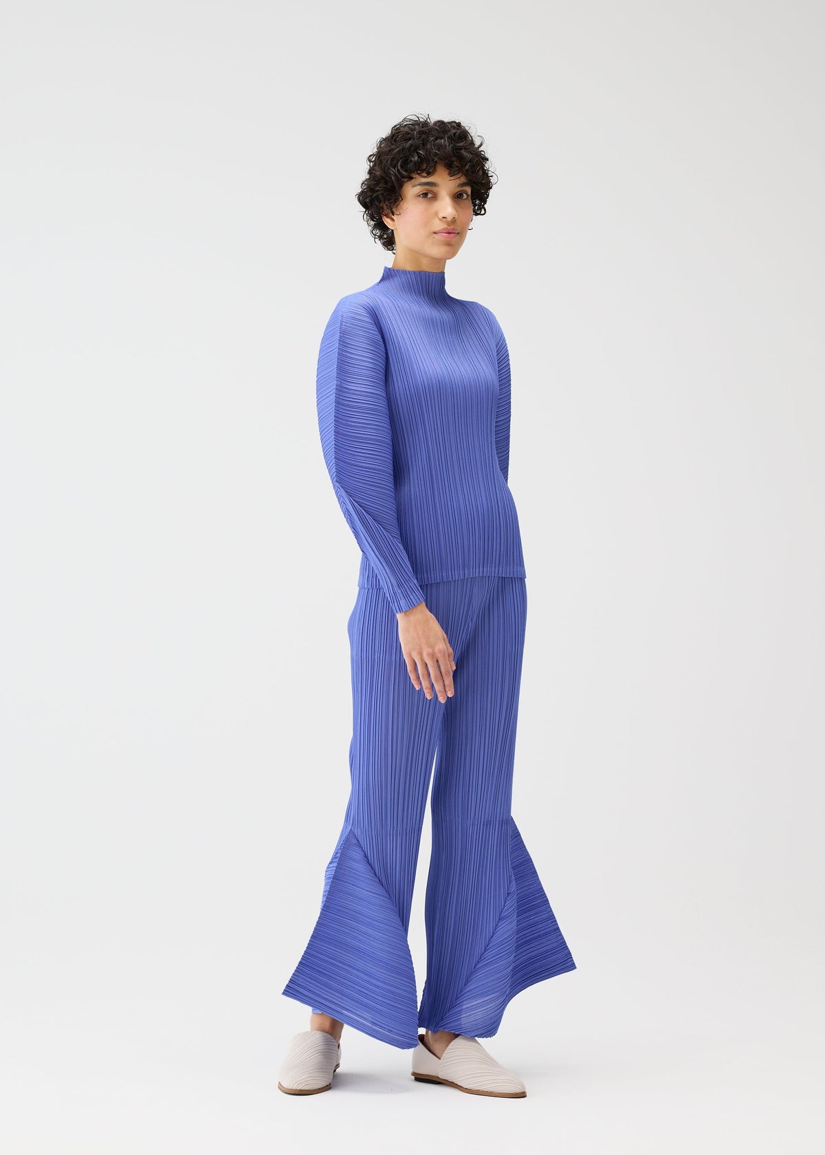 A person with short curly hair models the SKYLINE set by PLEATS PLEASE ISSEY MIYAKE: a textured blue long-sleeve top and flared city-inspired pants, styled with light slip-on shoes in a minimalist white studio. view 8 of 8